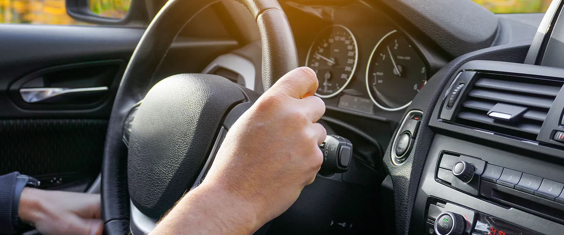 Driver's hands on a black steering wheel, dashboard visible. Sunlight.