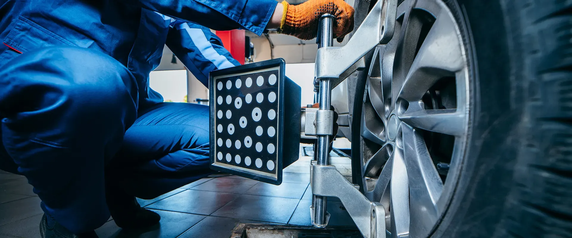 Mechanic in blue overalls, checking car wheel alignment with a digital tool.
