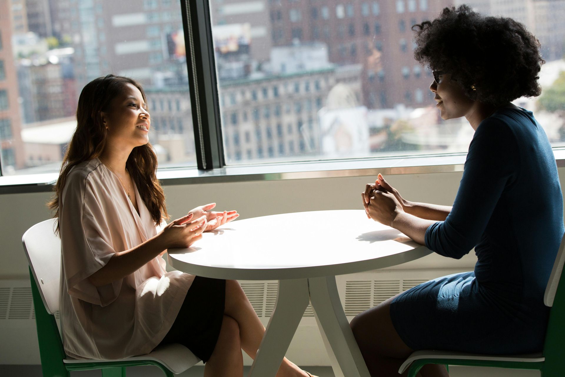 Two women seated at a round table near a large window are having a conversation in an office.
