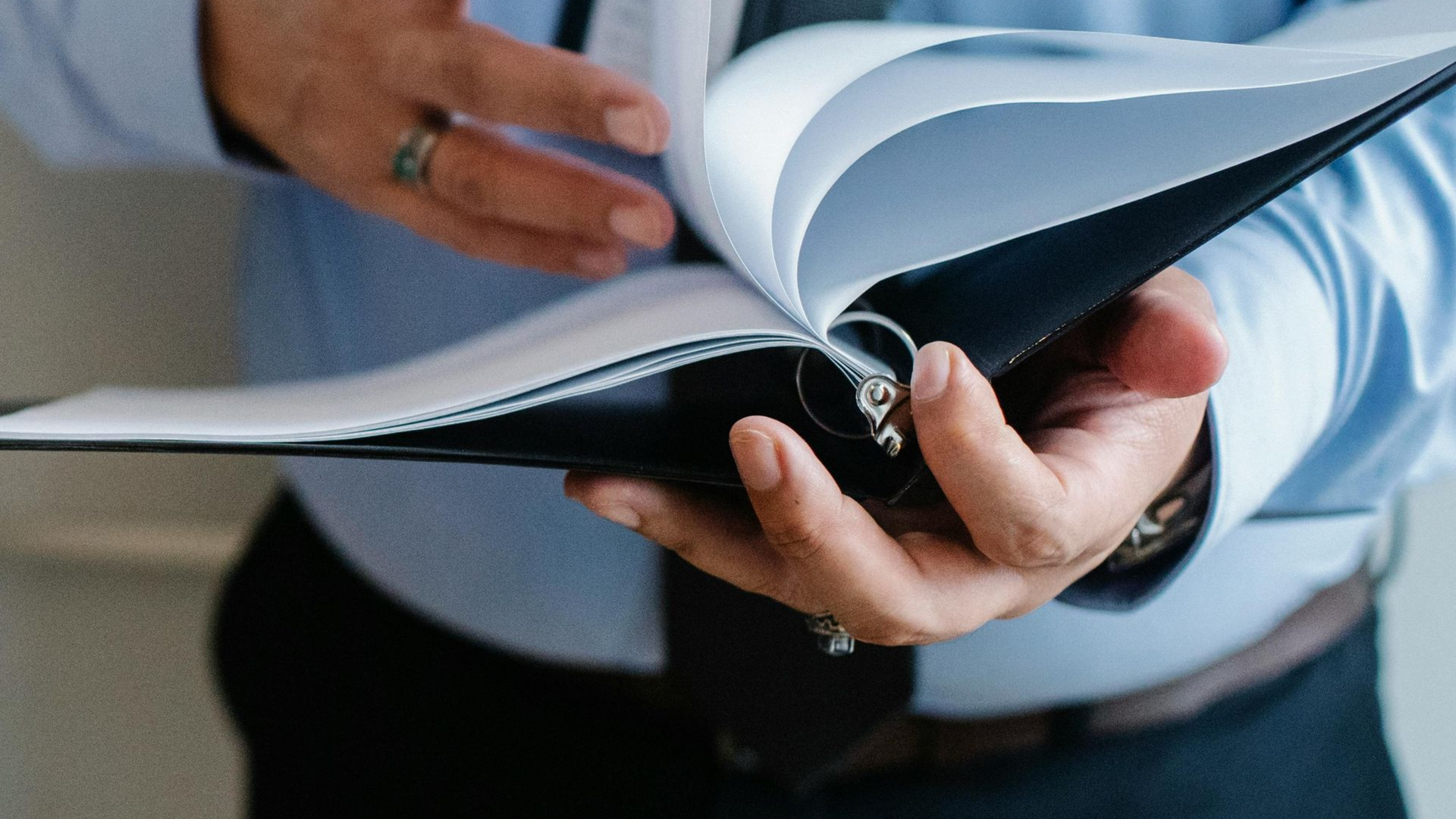 Person in a blue shirt with a black tie, holding and flipping through pages in a black binder.