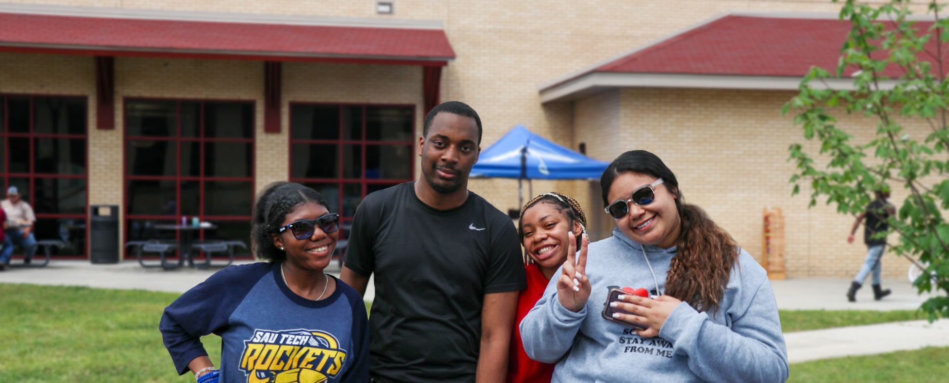 Four people pose for a photo in front of a building with red trim. One person makes a peace sign.