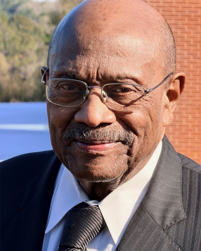 Older man with glasses, smiling, wearing a suit and tie, against a blurred outdoor background.