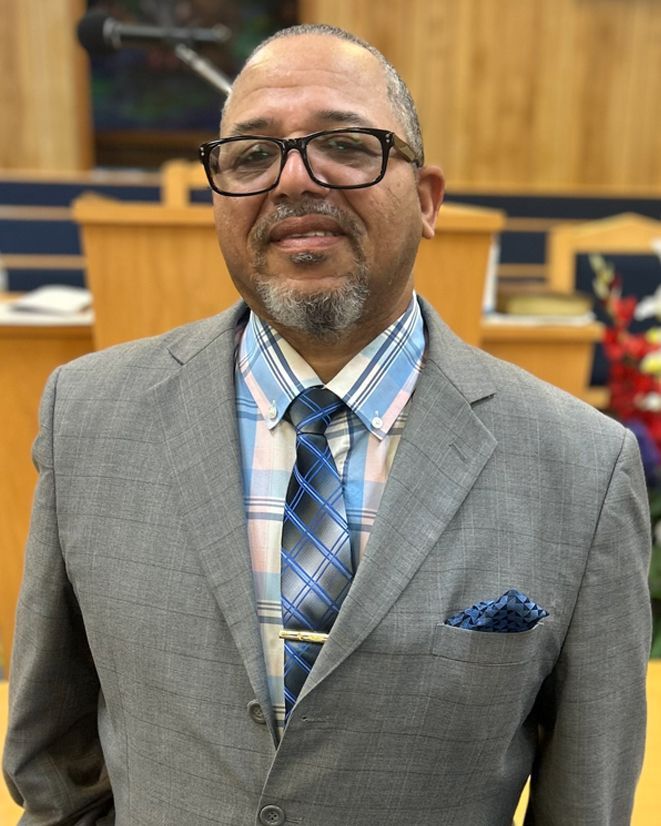 Man in glasses and suit, standing indoors, smiles at the camera. He is wearing a patterned tie and has a pocket square.