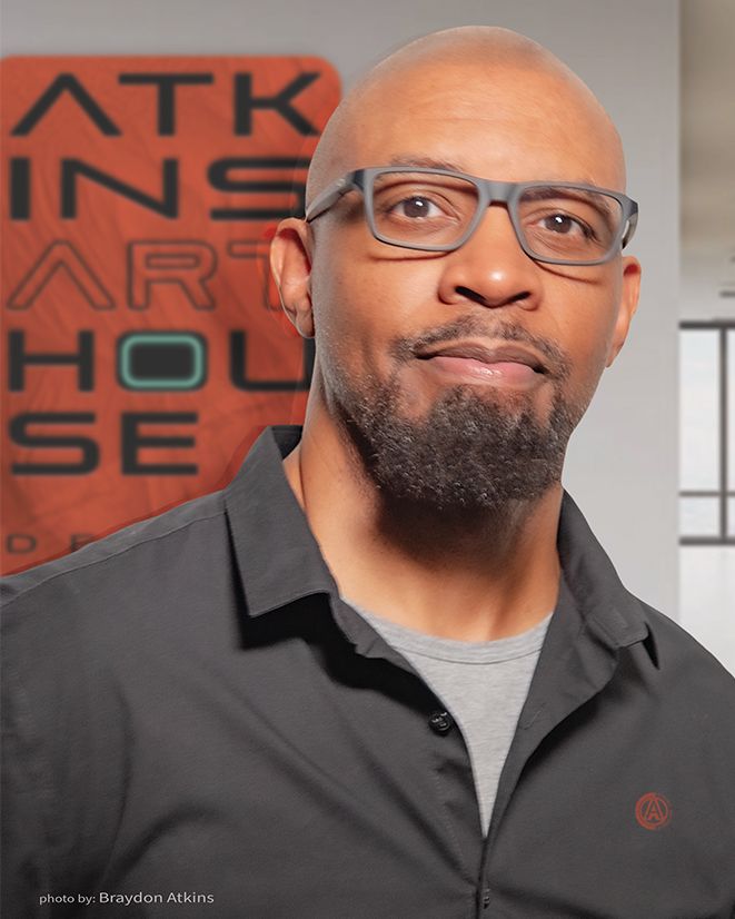 Man with a goatee and glasses, wearing a black shirt, in a room with a sign on the wall.