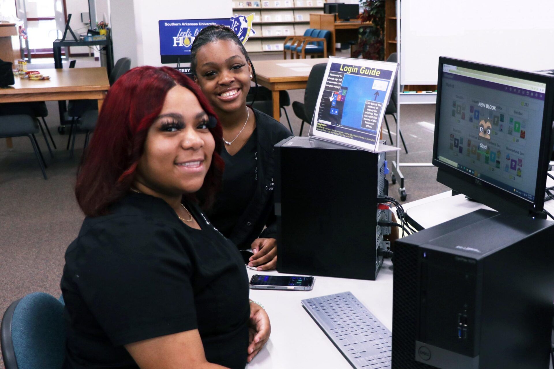 Two people at a computer in a library. One person with red hair smiles at the camera.