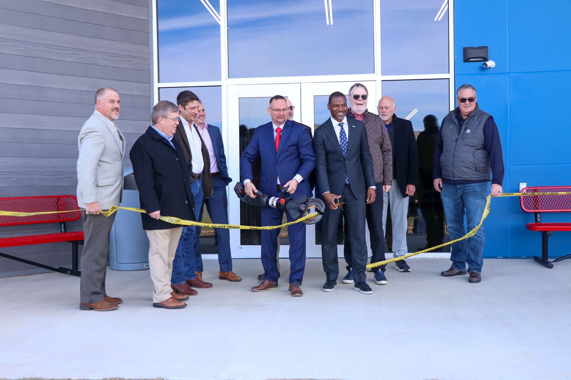 Group of men cutting a ribbon in front of a blue building, marking an opening.