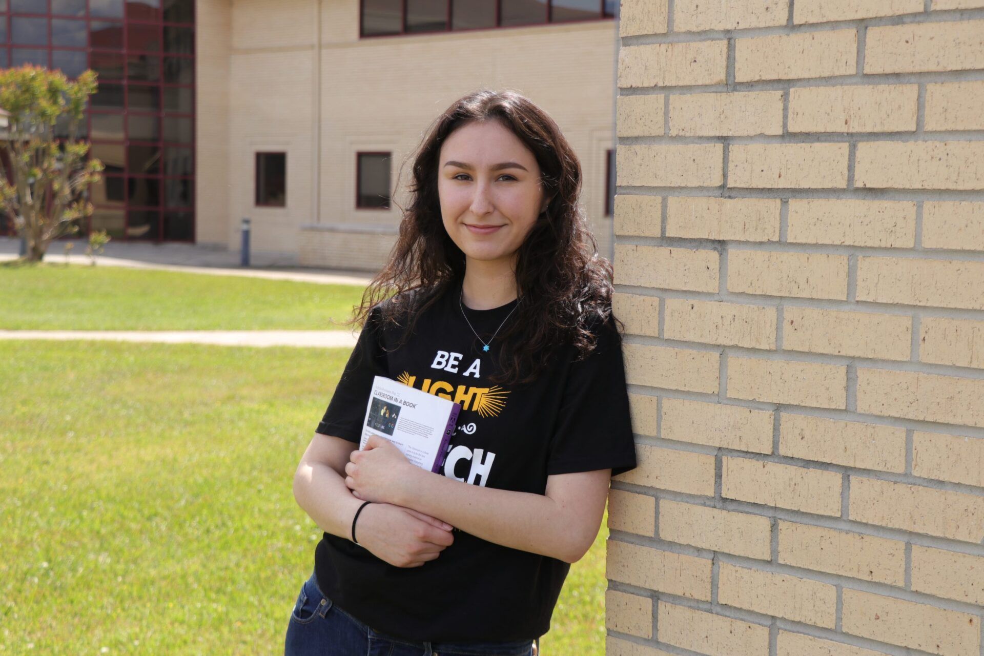 Person leaning against a brick wall, holding a book, smiling. Outdoors, near a building.