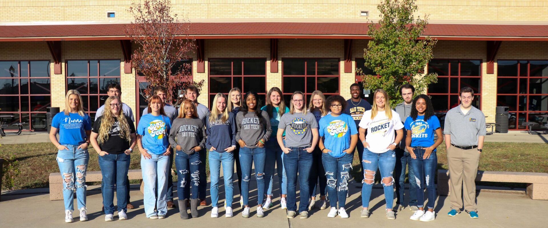 A group of students stand in front of a school building, smiling. They wear jeans and various t-shirts.