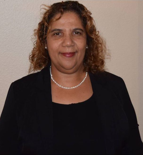 Woman with curly hair wearing a black jacket and a pearl necklace, smiling.