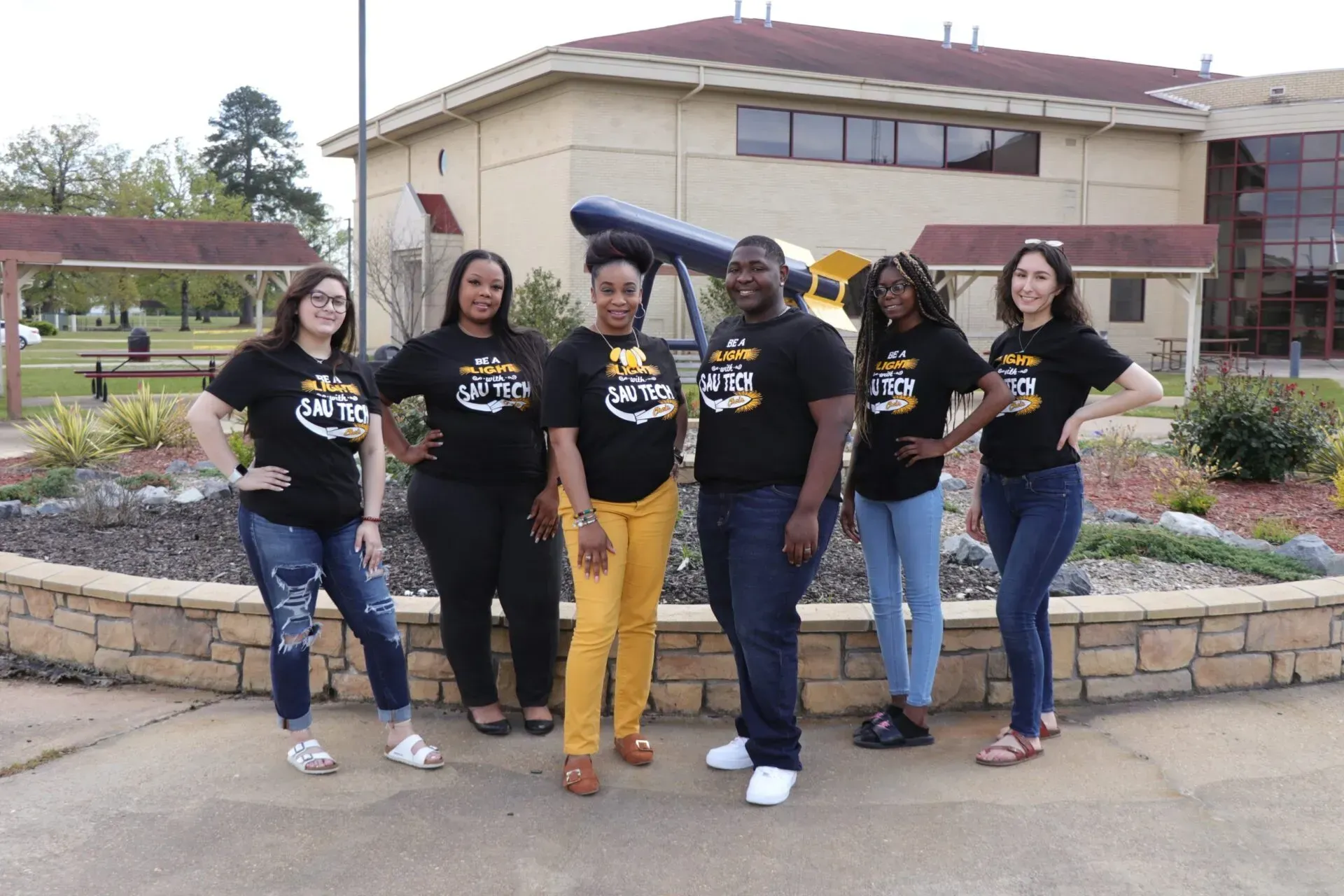 Six people pose outdoors in matching black shirts, in front of a building with a telescope.