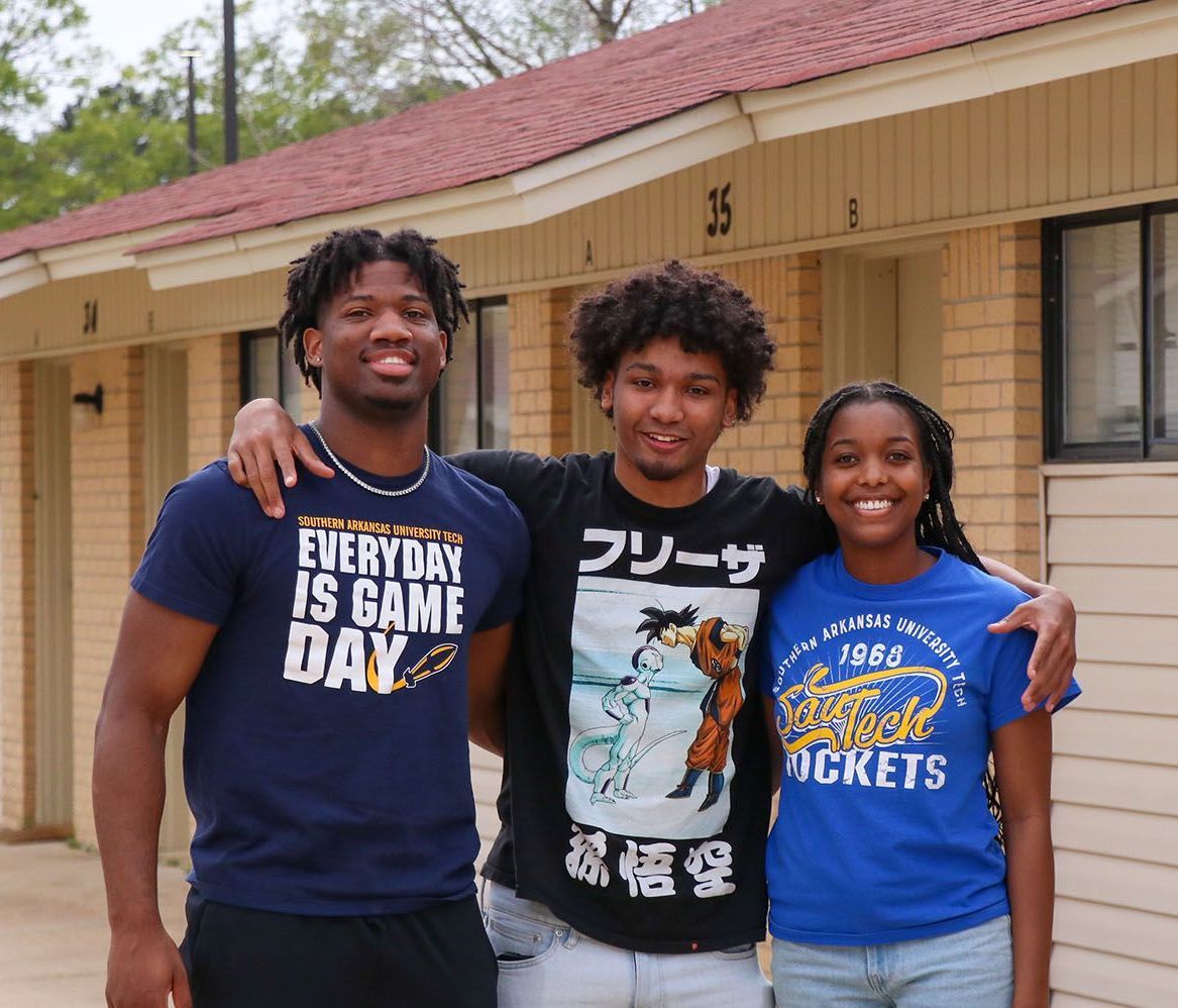 Three people smiling, posing in front of a building with doors. Two have arms around the middle person.