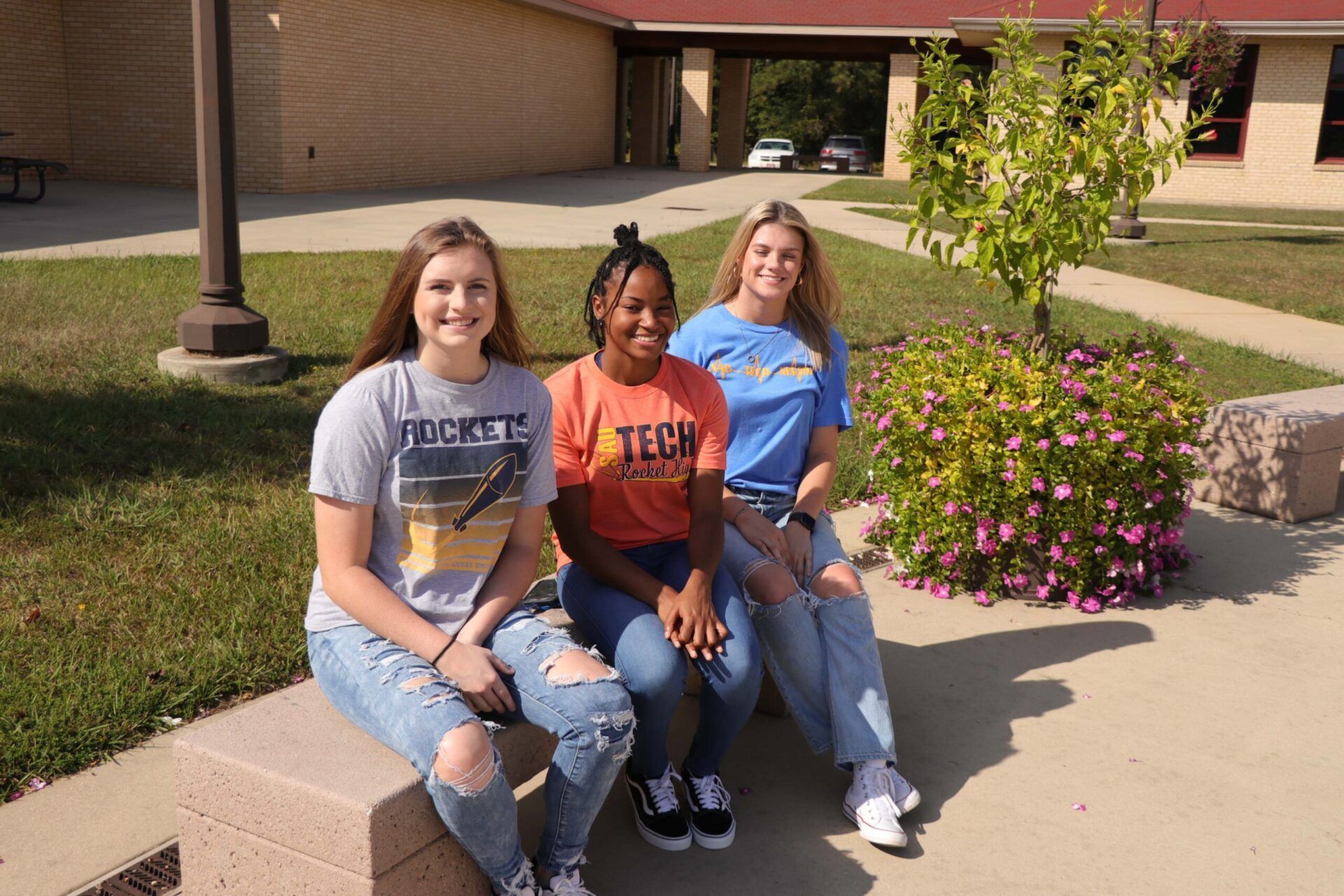 Three people in university shirts sit on a ledge outside; one has a gray shirt, one orange, one blue.