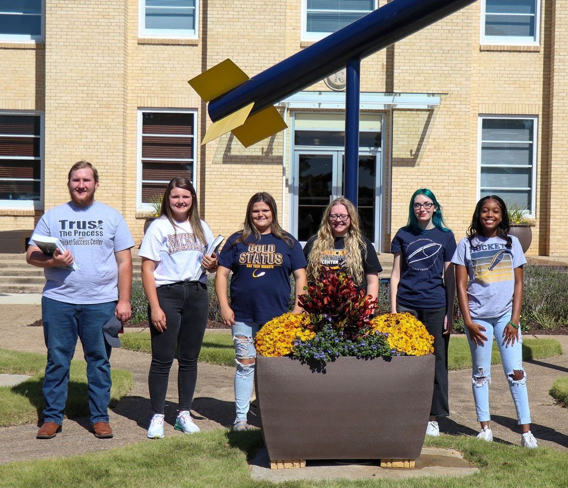 Six people stand in front of a brick building and large planter with a dark blue and yellow sculpture on top.