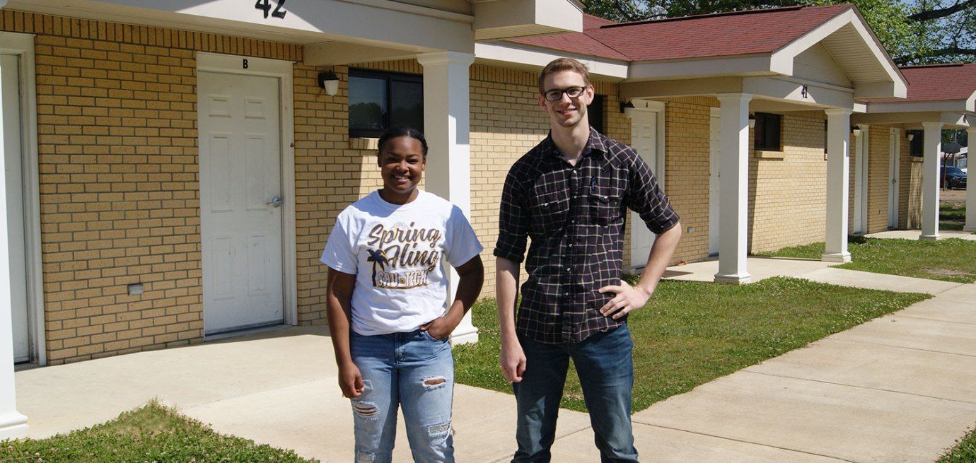 Two people standing in front of a brick apartment building. One wears a t-shirt and jeans, the other a plaid shirt and jeans.