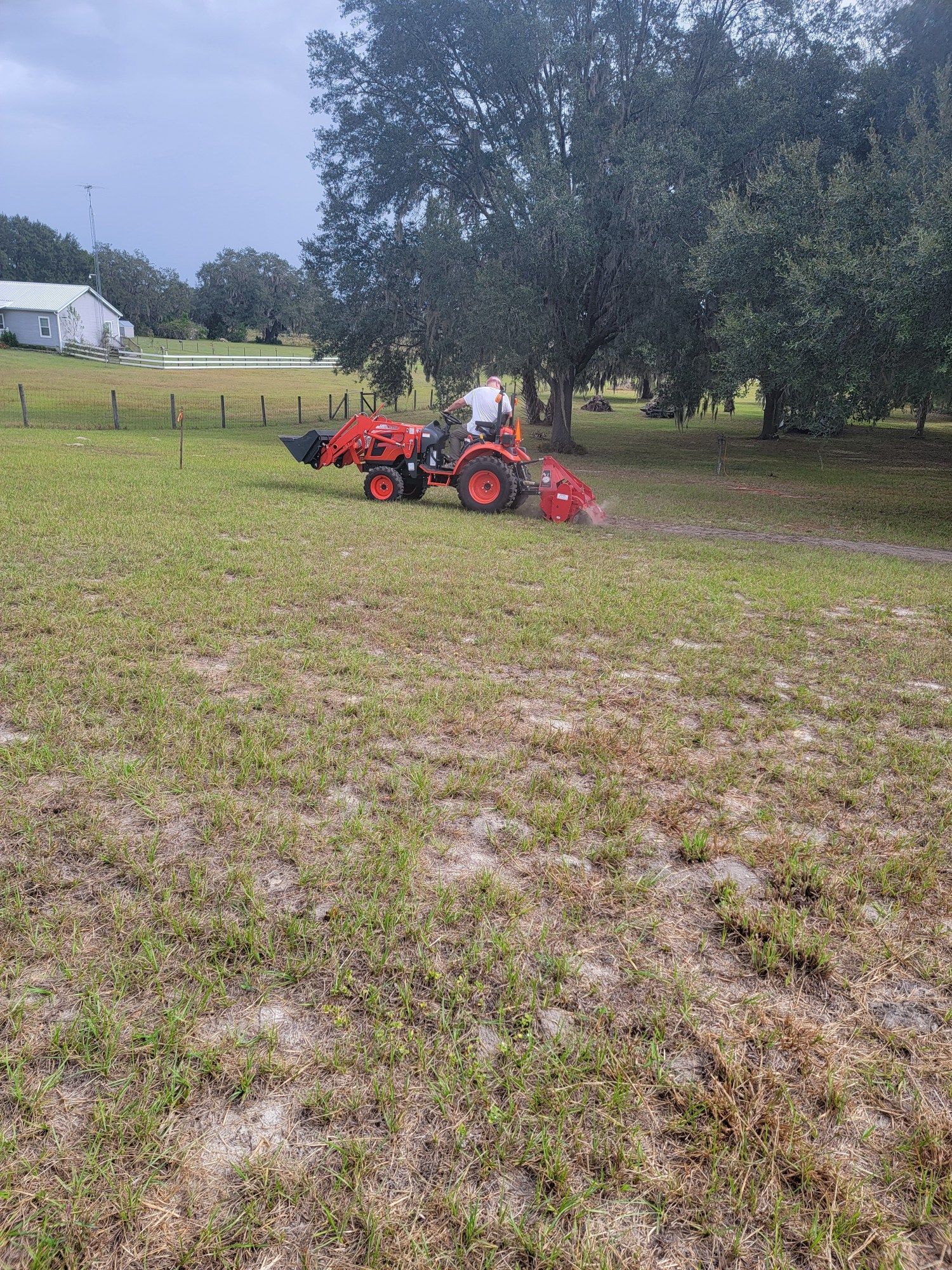 Person operating a red tractor with attachments on a grassy field, near trees and a fence.