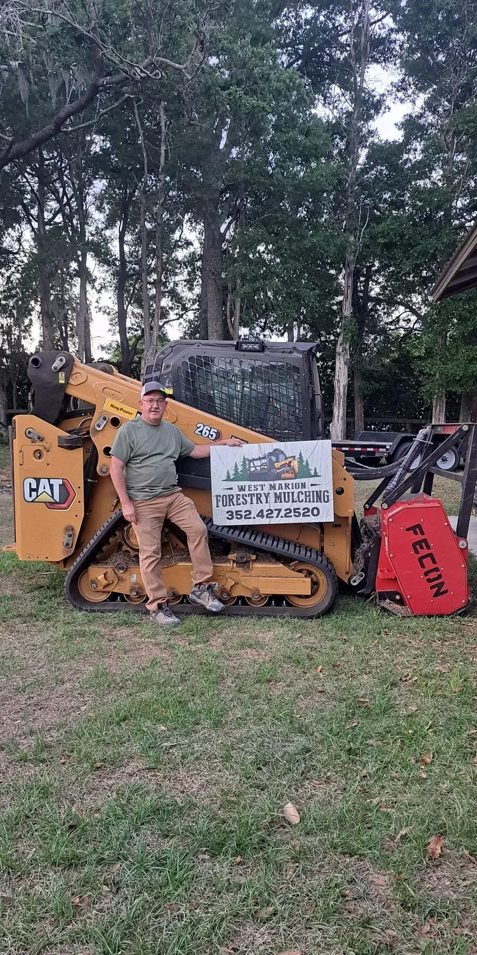 Man stands next to yellow CAT skid steer, holding sign with company logo on grass.