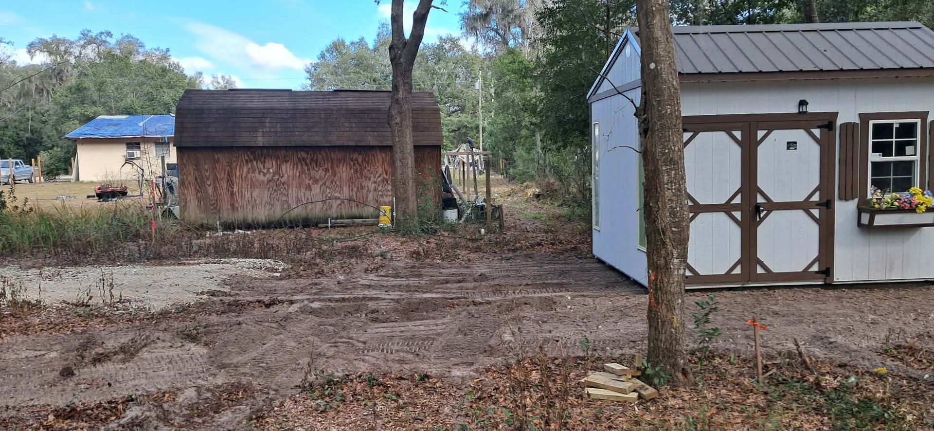 Rural yard with two small sheds, trees, and a white outbuilding in the foreground.