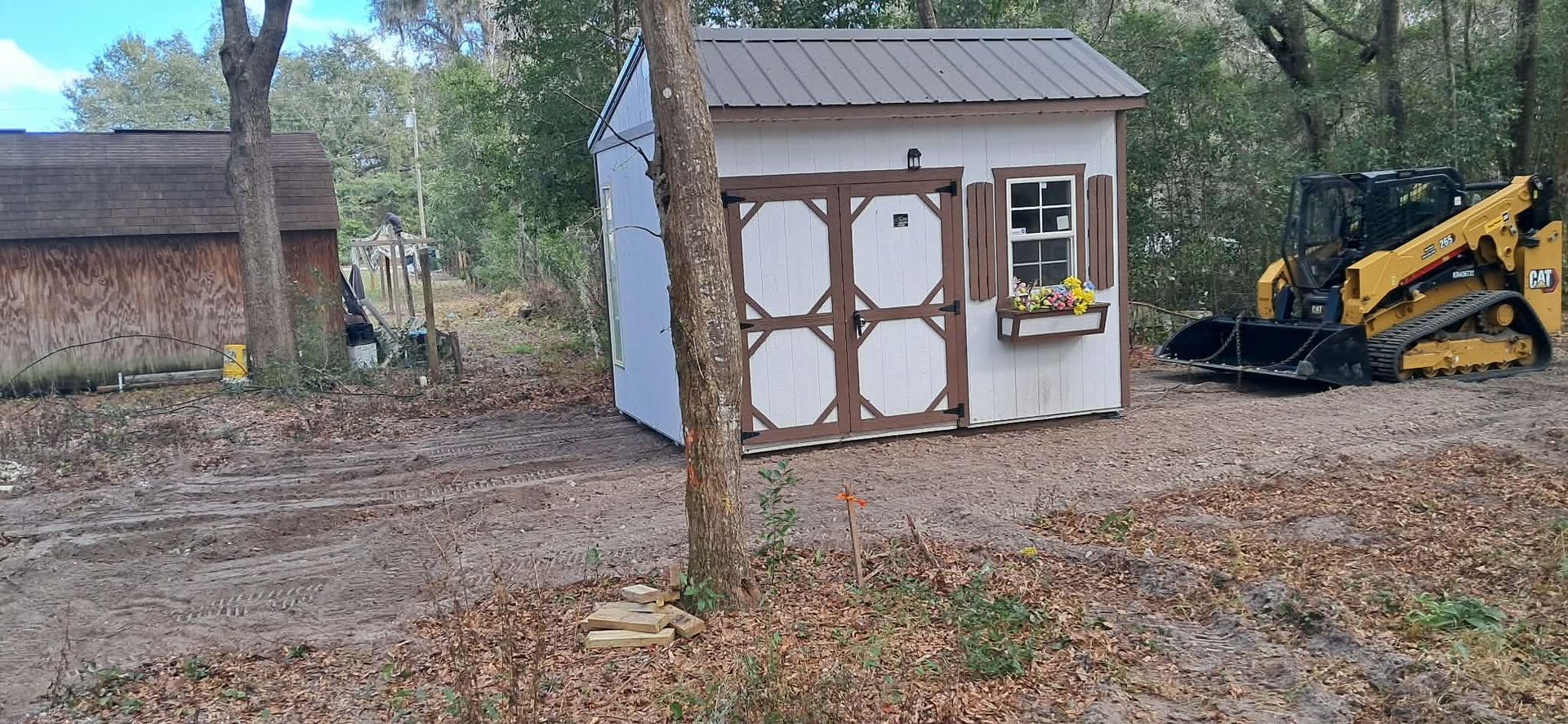 Backyard shed with a white door and tree in front, beside a yellow excavator on a leaf-covered ground