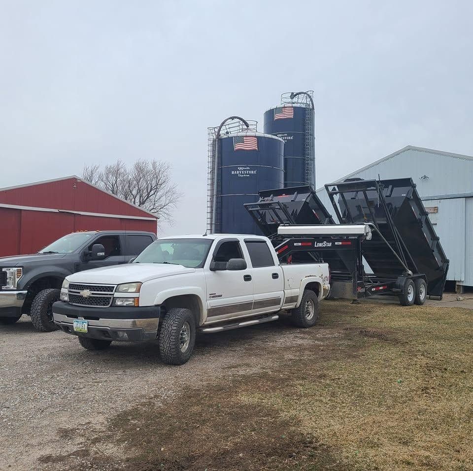 White pickup truck beside black dump trailer and large silos on a farmyard on a cloudy day