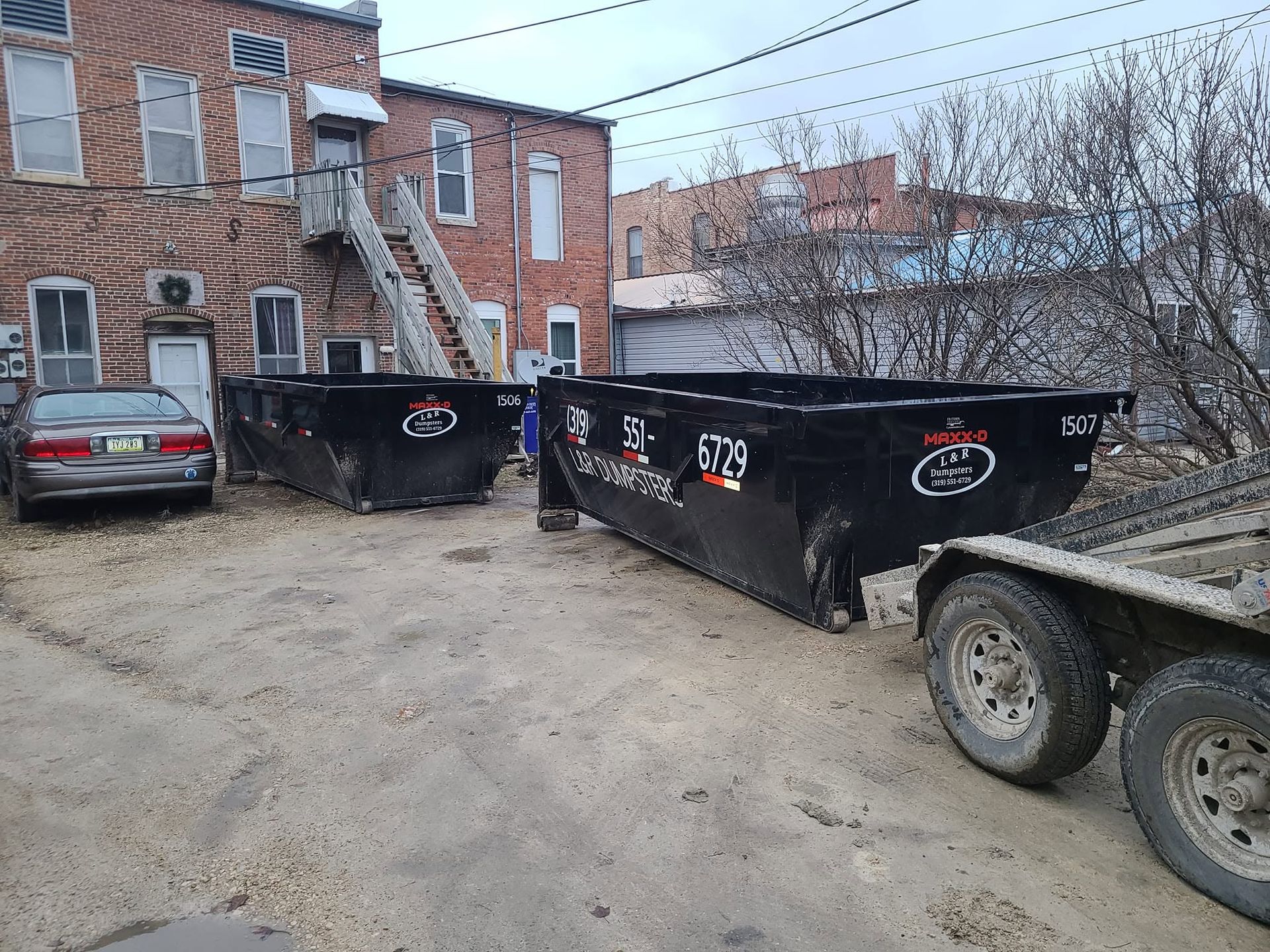 Dumpster and trailer in a brick apartment courtyard with parked cars and dirt ground