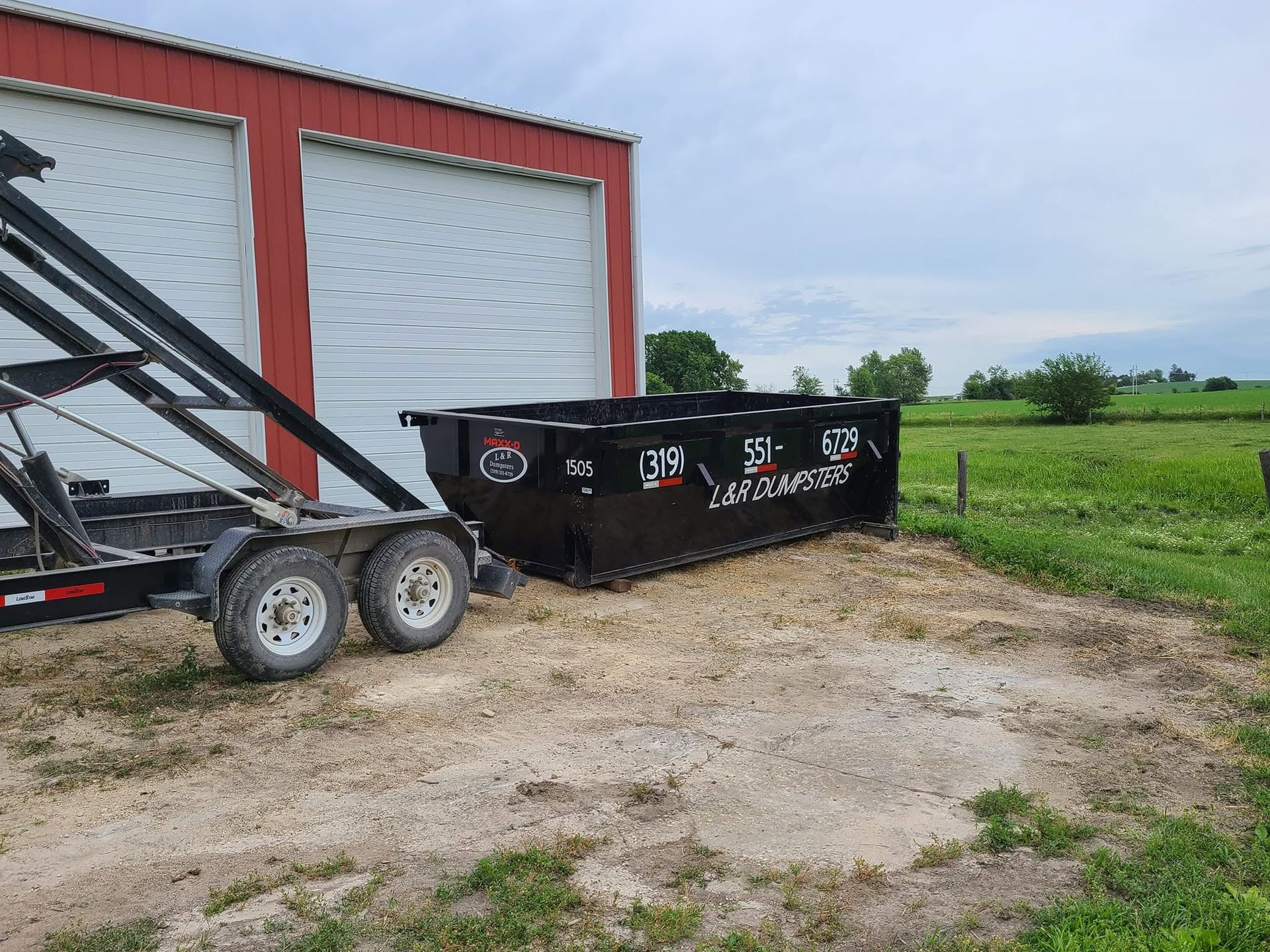 Black dumpster beside a red barn with white doors in a rural gravel lot