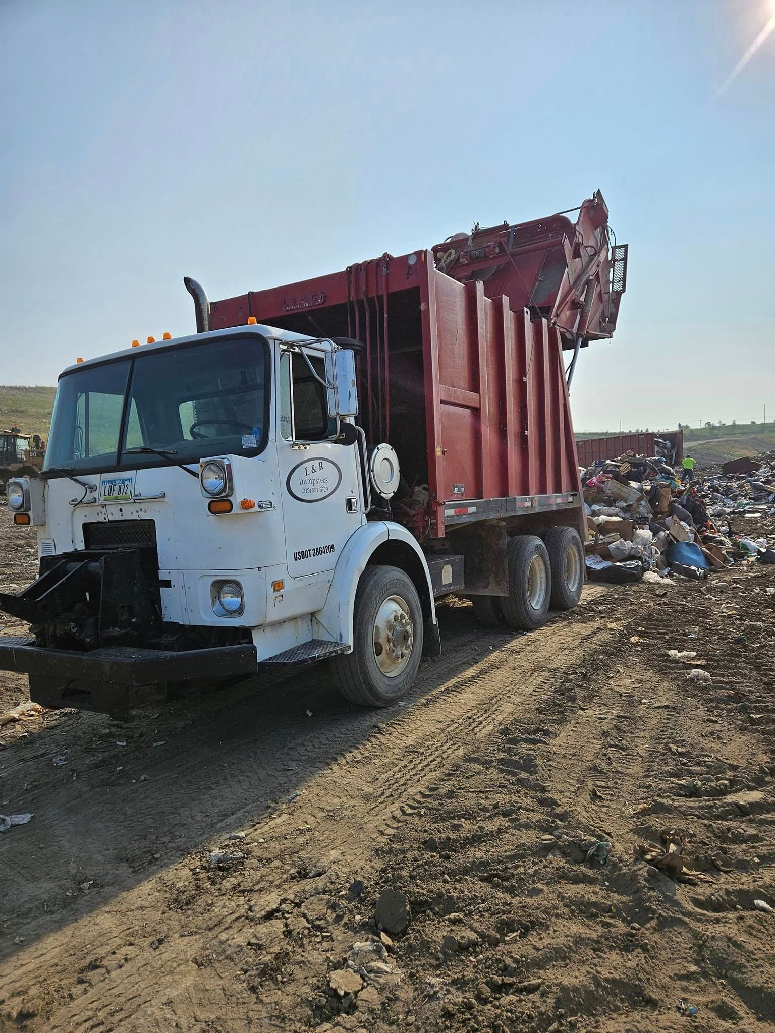 White and red garbage truck on a dirt road beside a large landfill under a bright sky