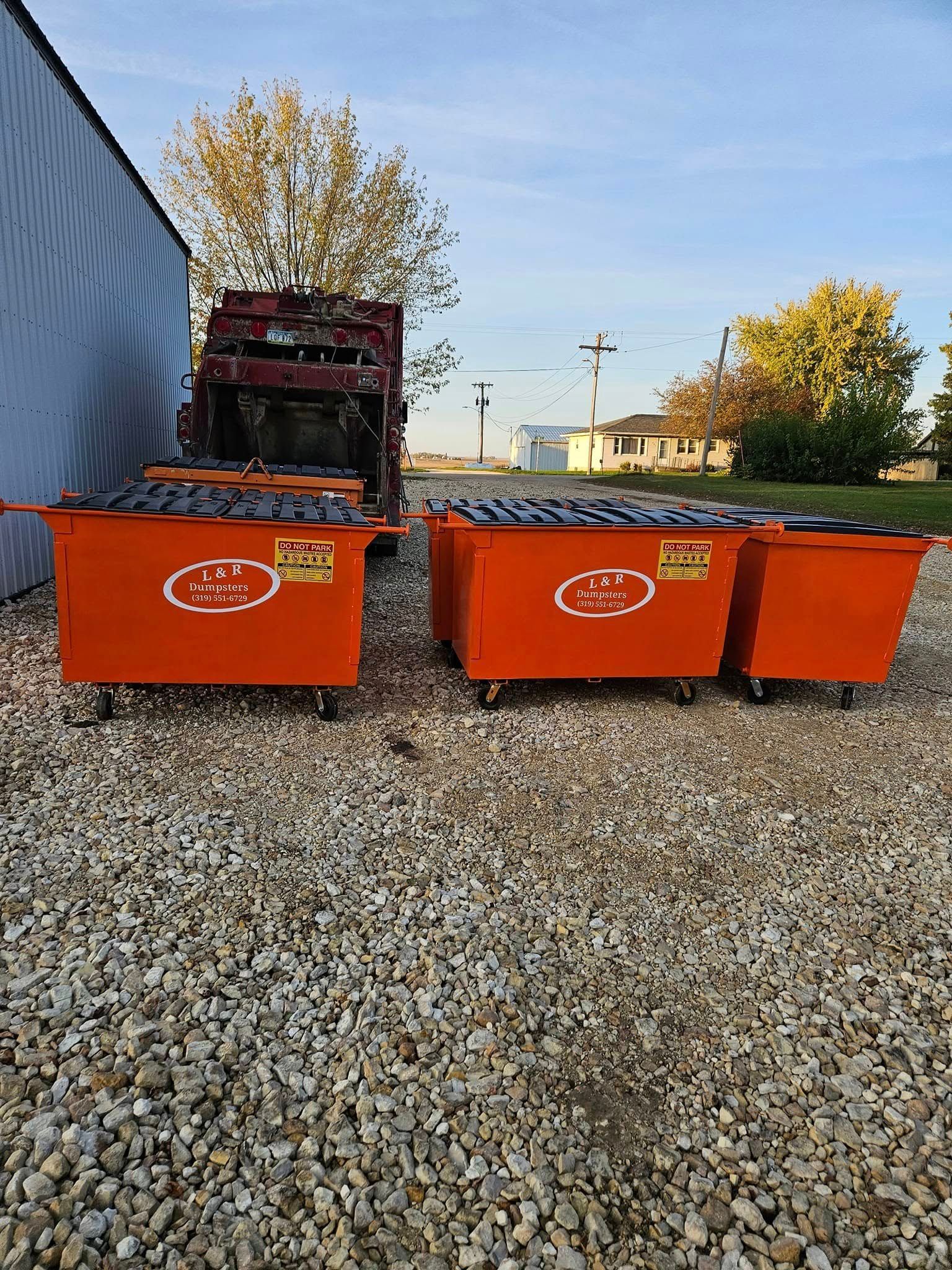 Three orange dumpsters on gravel beside a blue building, with trees and utility poles in the background.