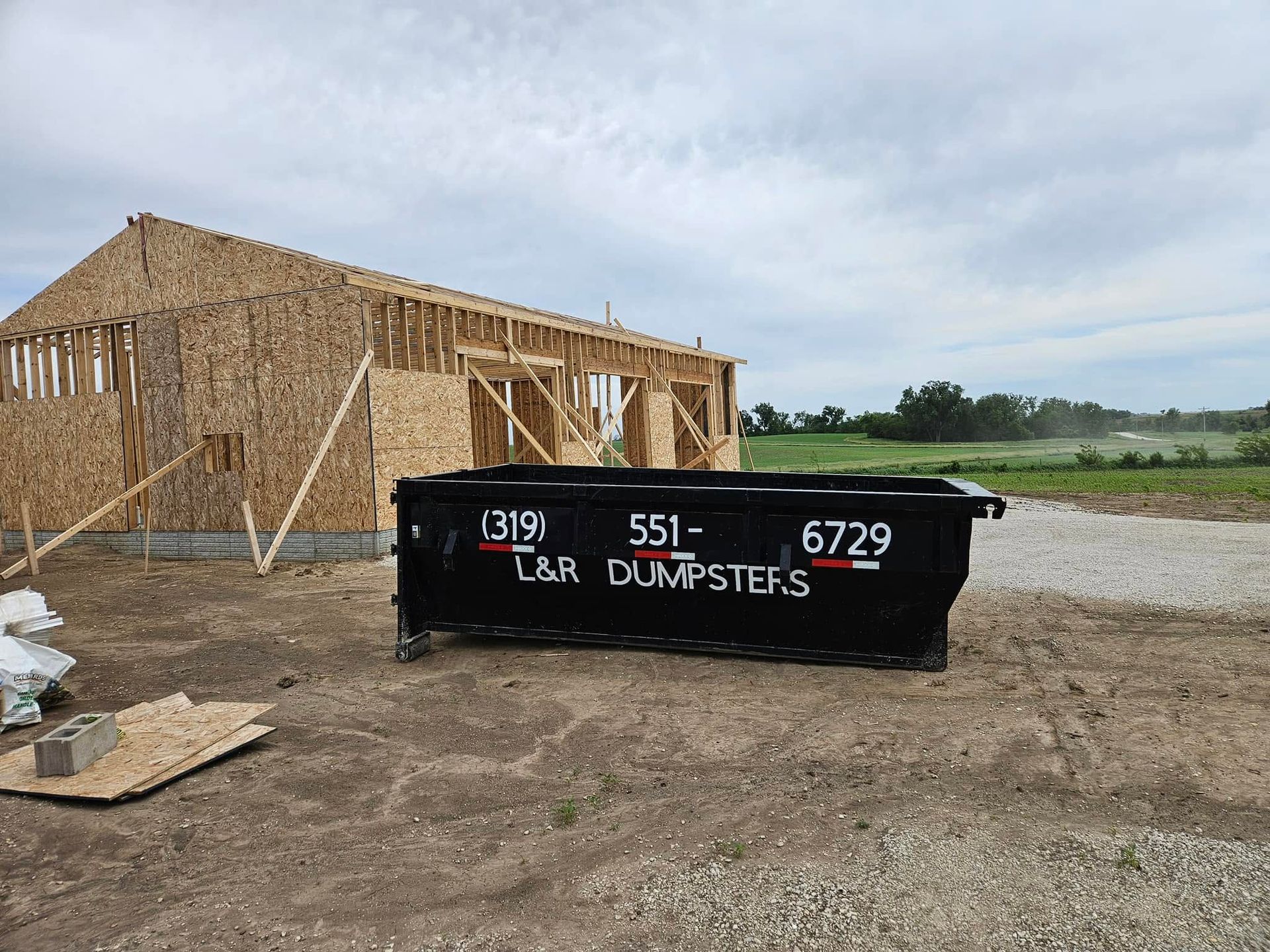 Black dumpster in front of a wood-framed house under construction on a dirt lot