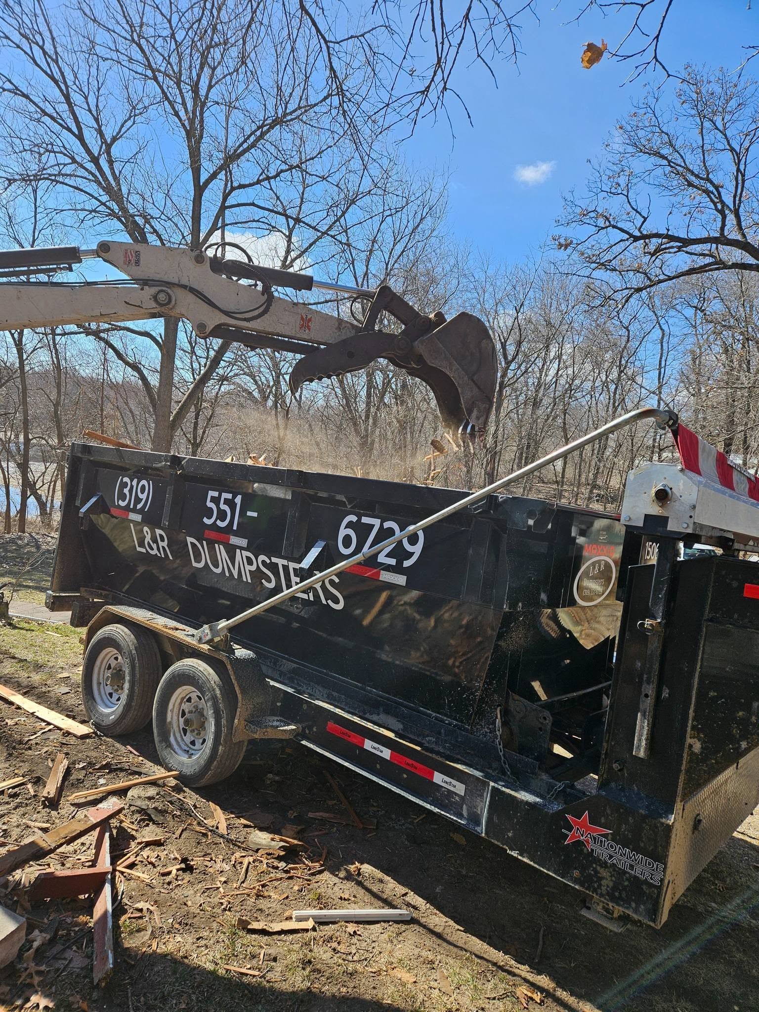 Black dump trailer with debris being loaded by an excavator in a wooded area