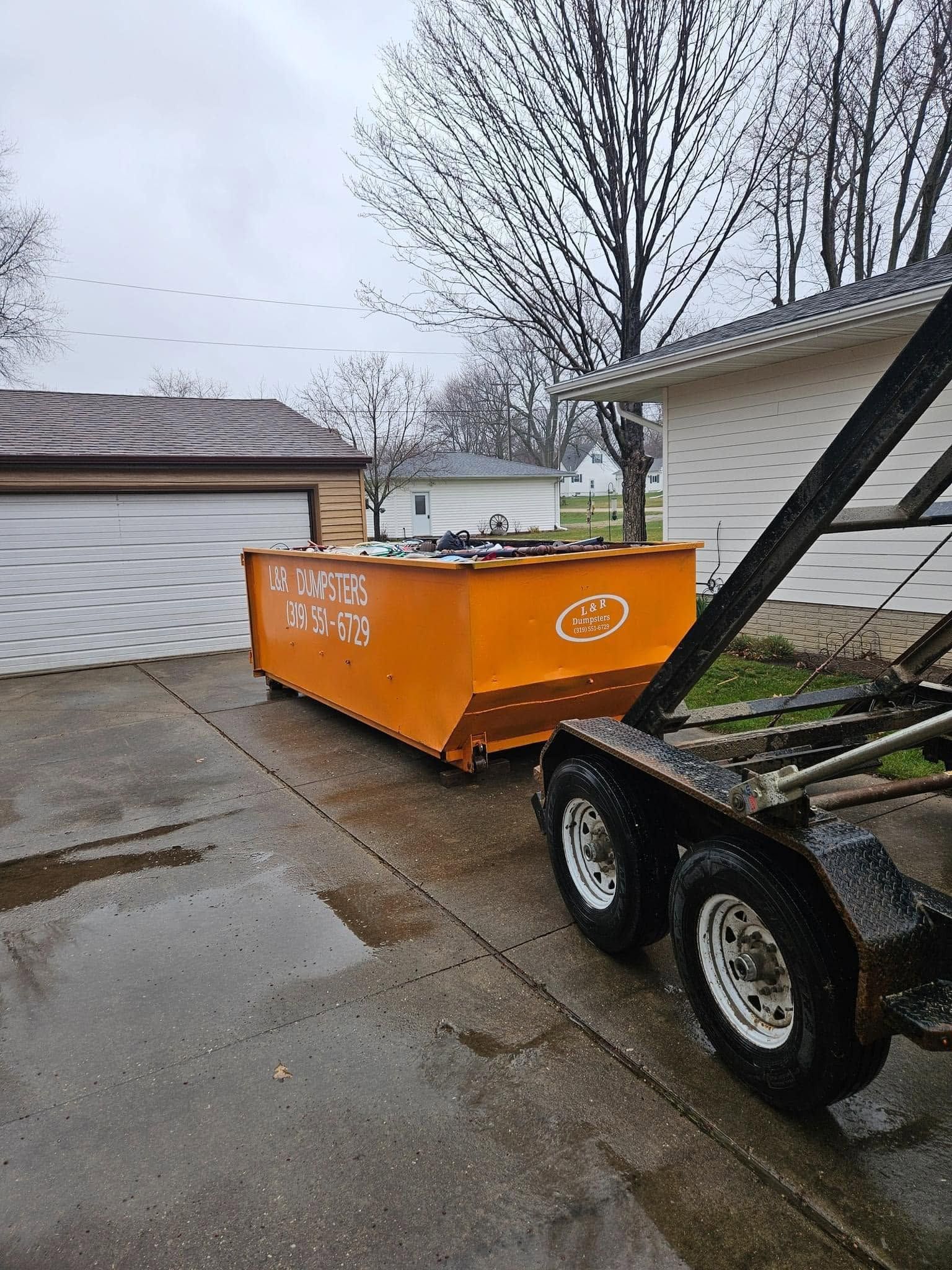 Orange dumpster on a trailer in a wet driveway beside a garage, with a leafless tree overhead.