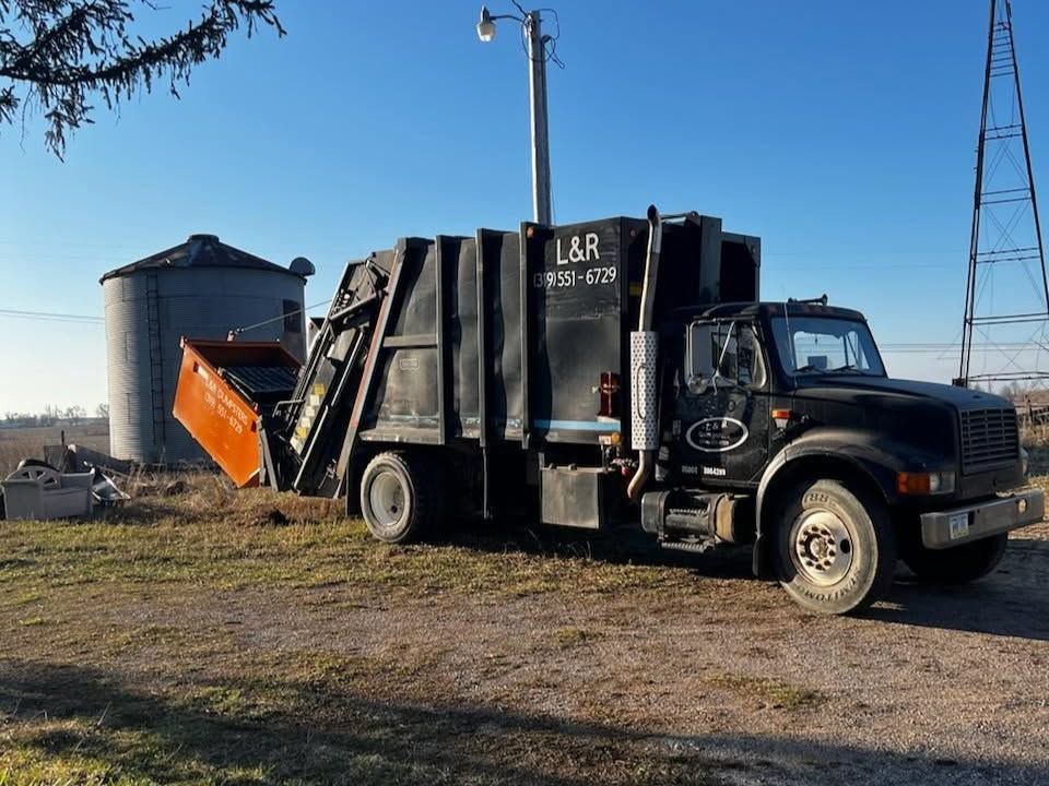 Black garbage truck with orange plow parked beside large storage bins on a gravel lot under clear sky