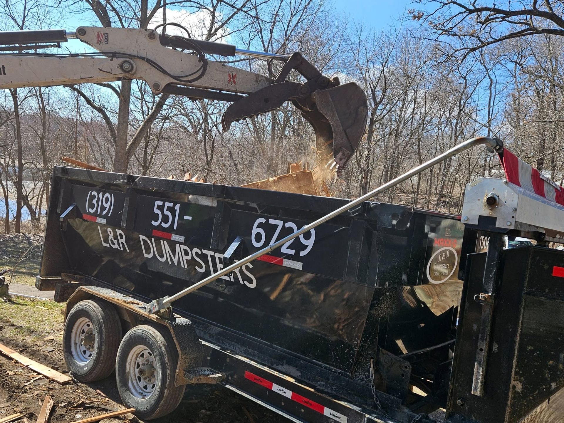Black dumpster trailer being loaded by an excavator in a wooded area
