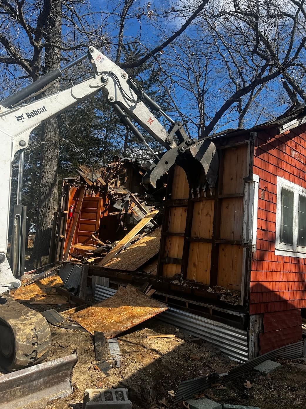 Excavator demolishing a house wall beside a red brick exterior, with debris and exposed wooden framing.