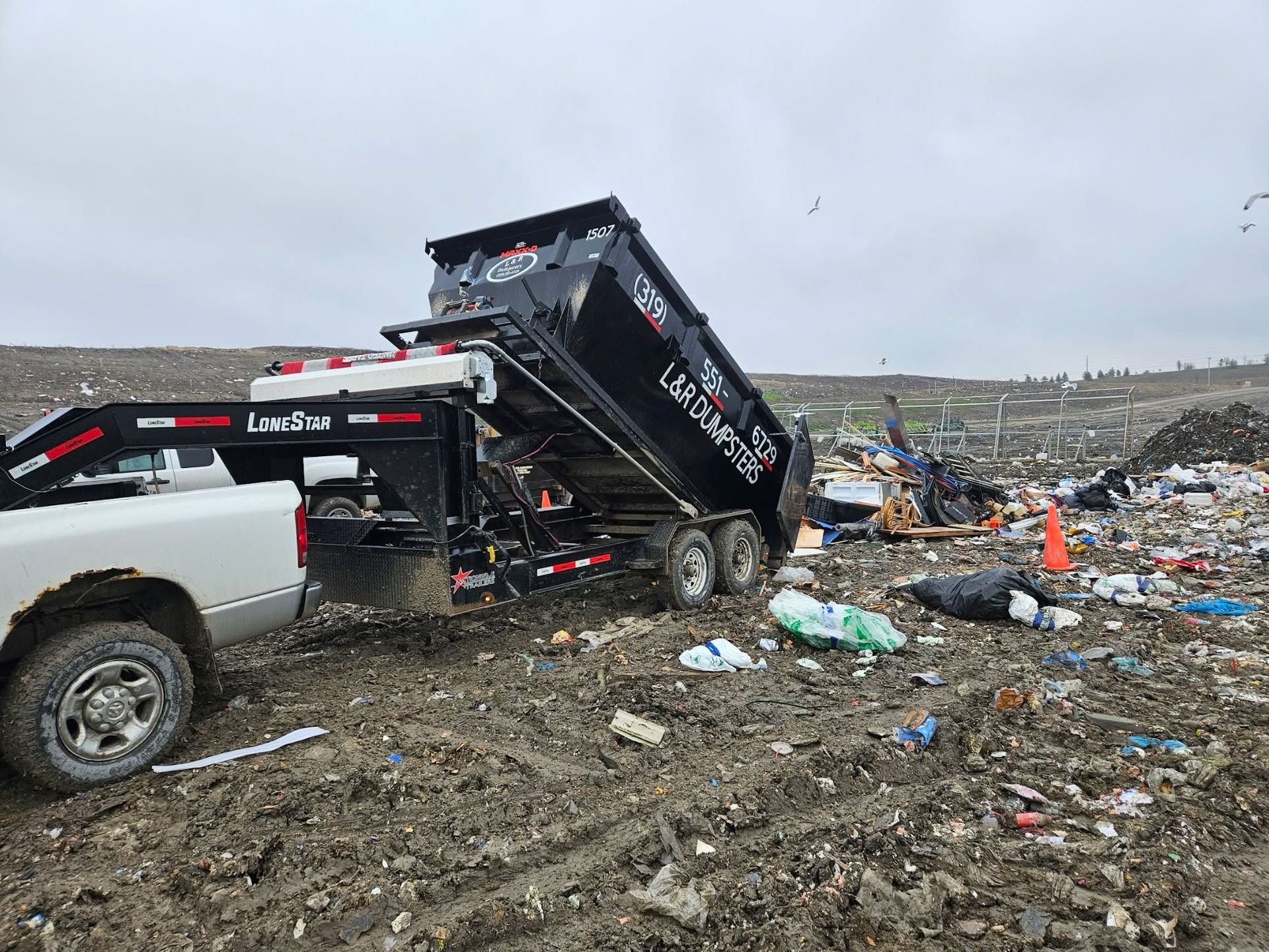 White dump truck unloading trash in a muddy landfill under a gray sky.