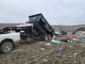 Tow truck dumping debris at a muddy landfill, with scattered trash and an orange cone nearby