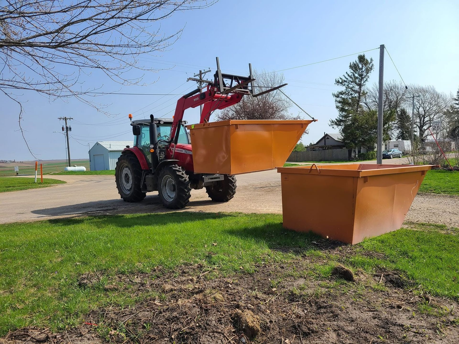 Red tractor with front bucket beside a large orange dumpster on a grassy roadside.