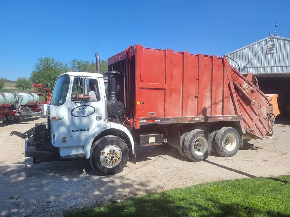 Red garbage truck parked on a gravel lot beside a barn under a clear blue sky