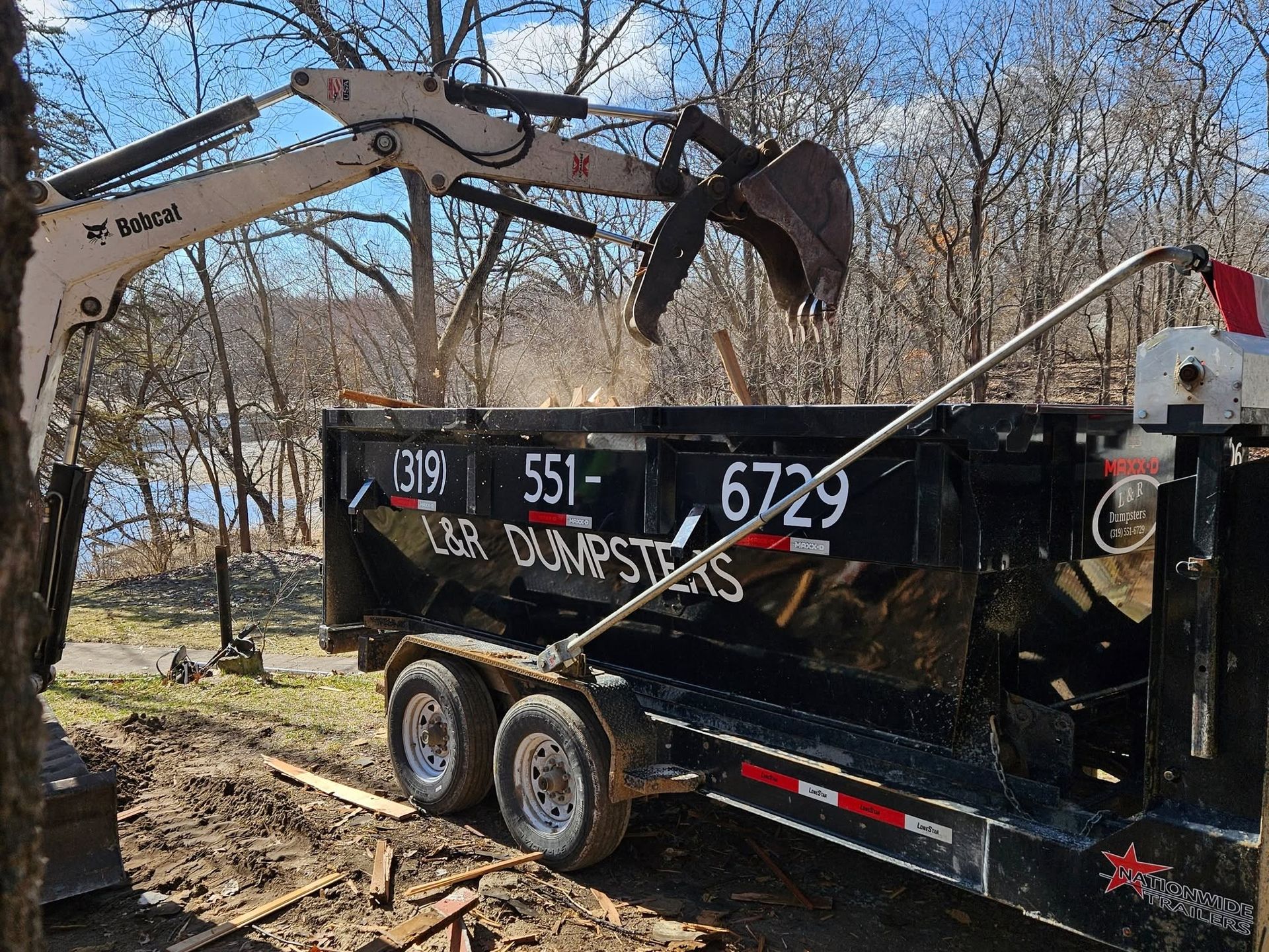 Excavator loading debris into a black dump trailer in a wooded area