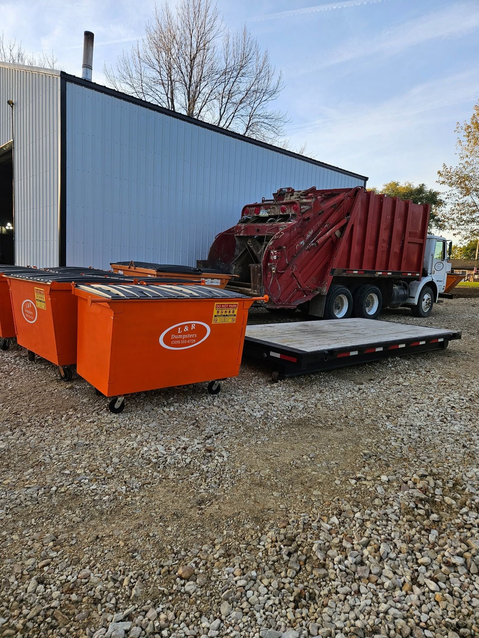 Red dump truck beside orange dumpsters on a trailer in a gravel lot near a metal building.