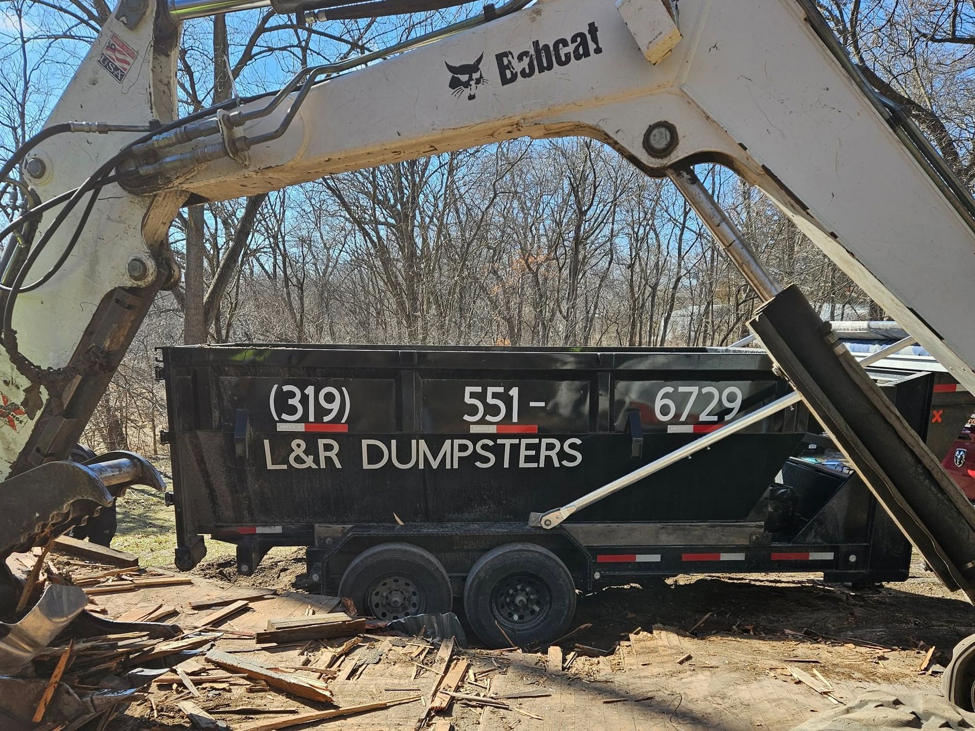 White Bobcat excavator dumping dirt into a black L&R Dumpsters trailer in a wooded lot