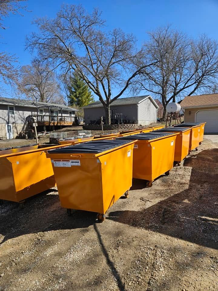 Row of bright yellow dumpsters in a yard beside buildings and bare trees on a sunny day