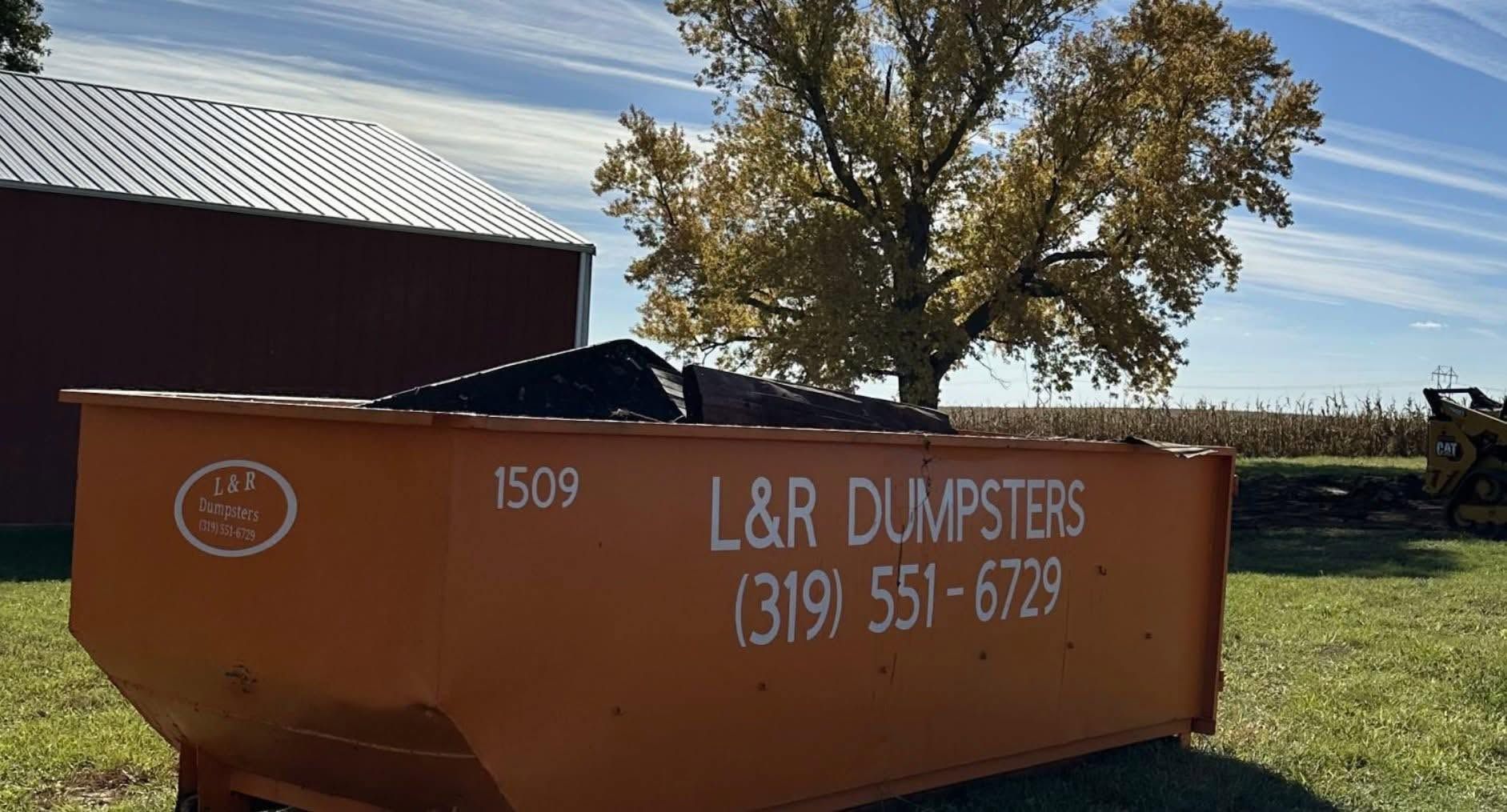 Orange L&R dumpsters beside a barn in a rural field under a blue sky