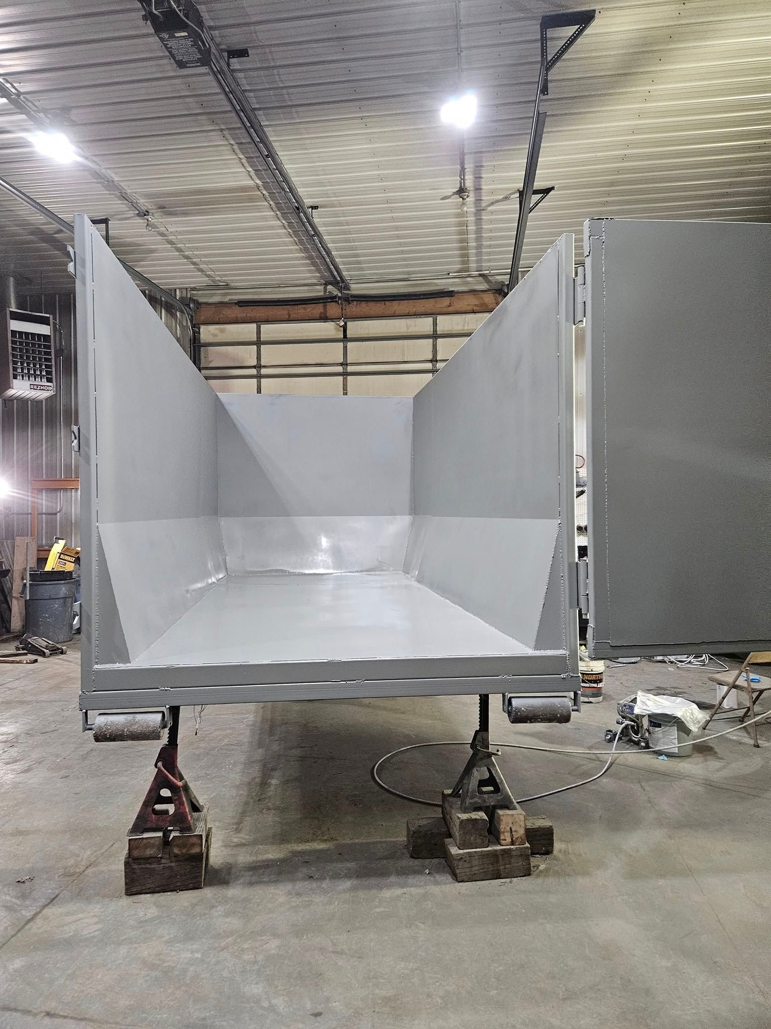 Gray industrial metal hopper trailer in a warehouse, propped on jack stands with its front raised.