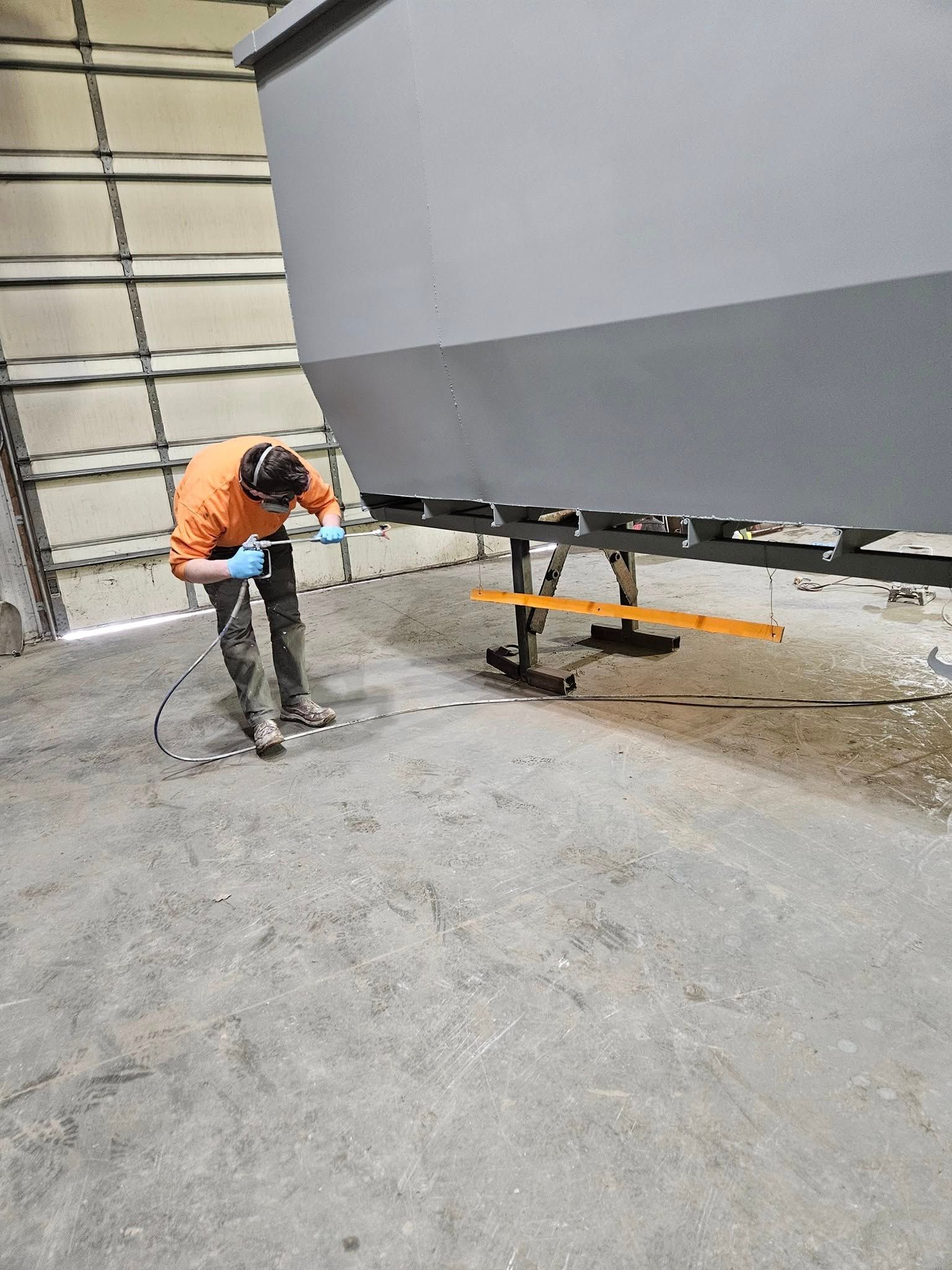 Worker in orange safety gear operating a power tool beside a large gray industrial structure.