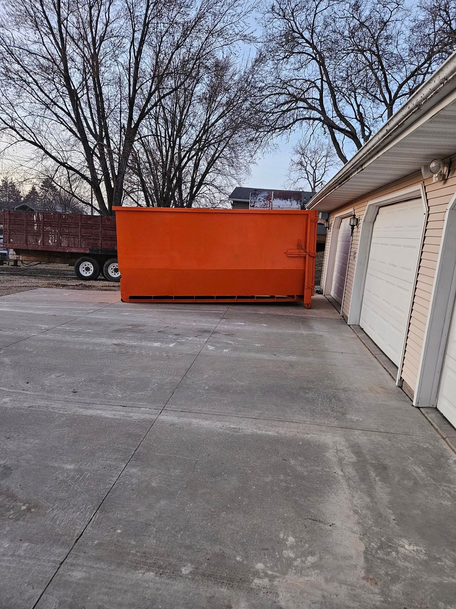 Orange dumpster in a concrete driveway beside a garage, with leafless trees in the background.