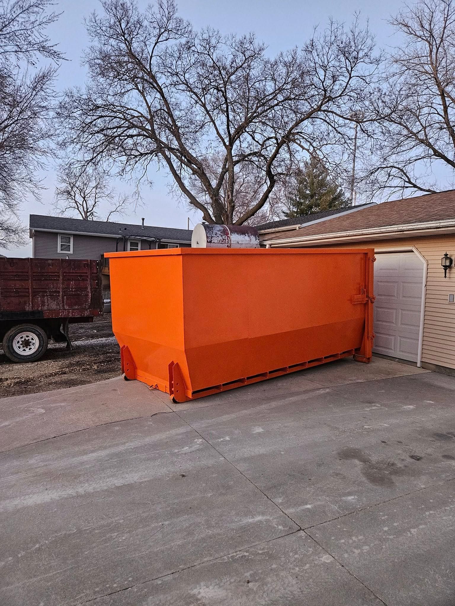 Orange dumpster in a driveway beside a beige garage, with a parked truck and trees in the background