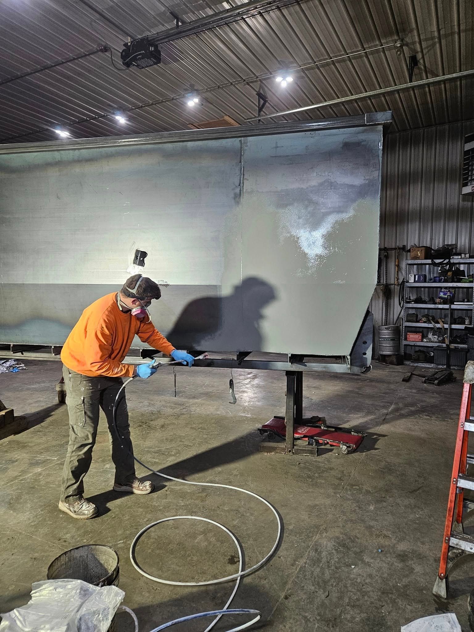 Worker in orange shirt sanding a large panel in a workshop, with tools and ladder nearby.