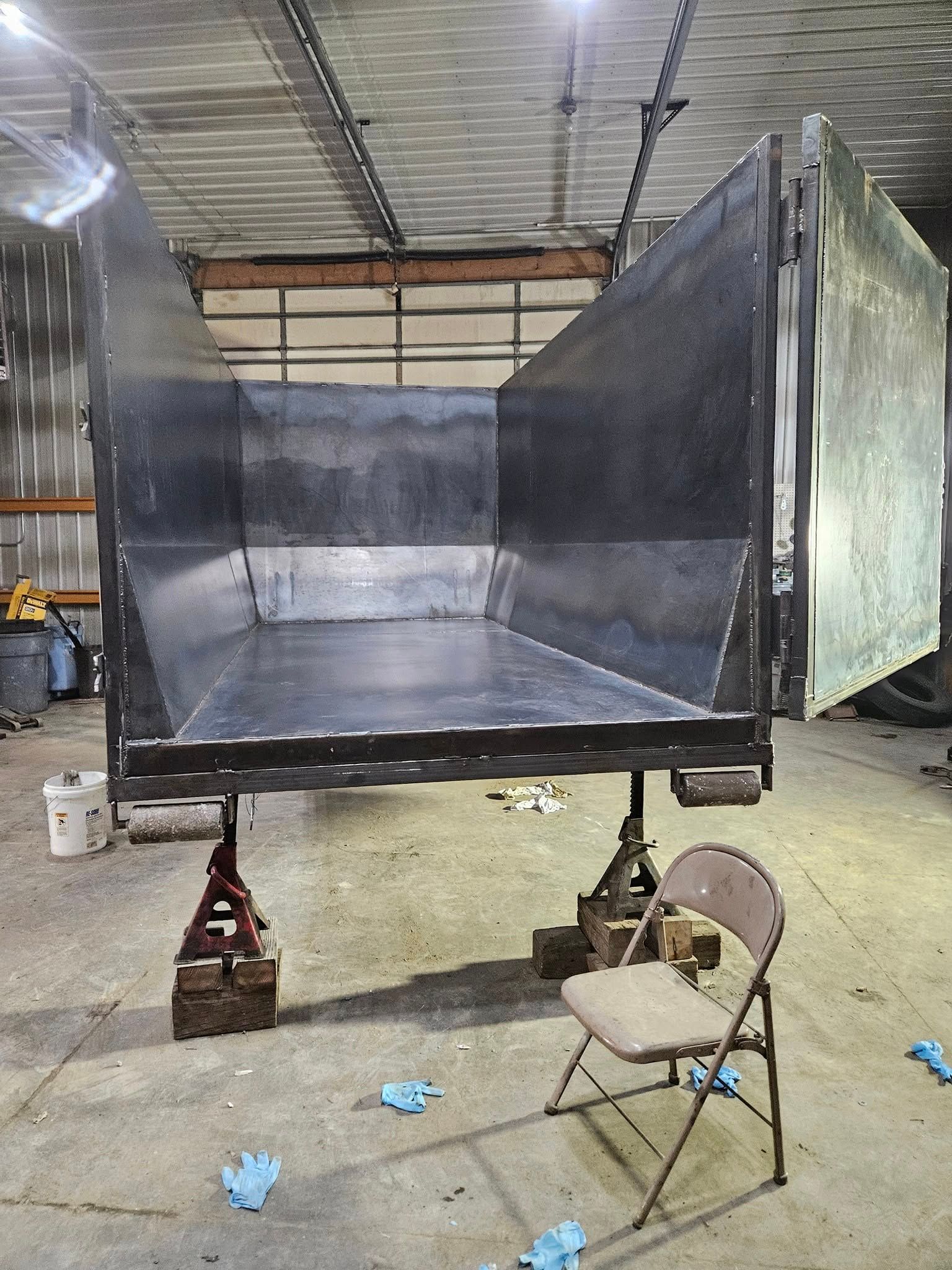 Large black metal container or trailer in a workshop, with a chair and jack stands underneath.