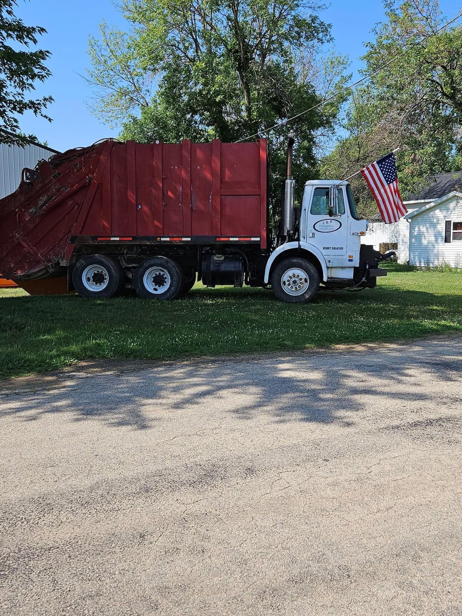 Red dump truck parked on grass beside a gravel drive, with trees and a house in the background.