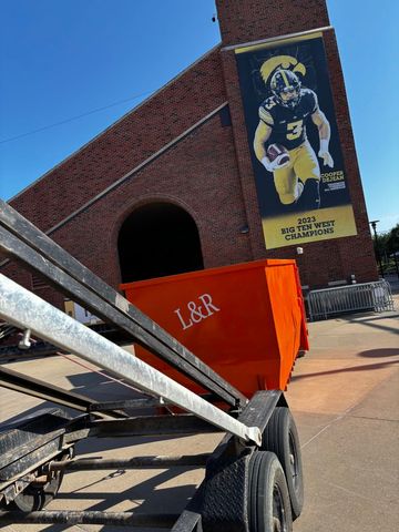 Orange dumpster on a wheeled cart in front of a brick building with a mural.